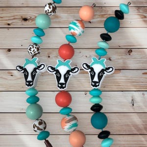 May include: Three beaded necklaces with cow-shaped pendants. The necklaces feature a variety of colourful beads, including turquoise, coral, black, and patterned beads. The cow pendants are black and white with turquoise accents.