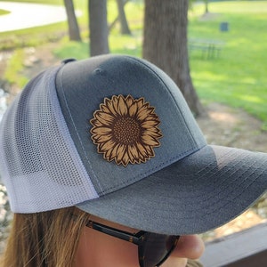 May include: A grey and white trucker hat with a brown leather sunflower patch. The hat is being worn by a person with brown hair and sunglasses.