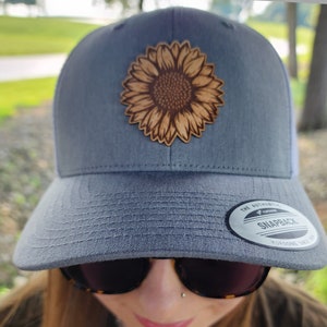 May include: A gray and white baseball cap with a brown leather sunflower patch on the front. The cap has a black and white "The Authentic Yupoong Snapback Yupoong Since 1974" label on the back.