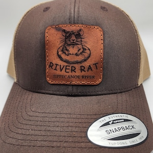 May include: A brown and tan trucker hat with a leather patch featuring a rat wearing sunglasses in an inner tube. The patch reads "River Rat Tippecanoe River." The hat has a snapback closure and a "Yupoong" tag.