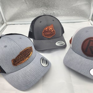 May include: Three gray and black trucker hats with different leather patches. The patches feature a logo with the text "Cardinal", a logo with the text "Lourasch Family Farms", and a logo with the letter "R".