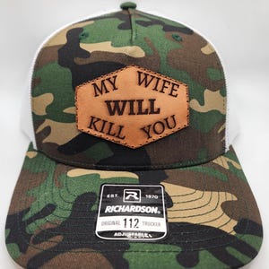 May include: Camouflage trucker hat with a white mesh back and a brown leather patch. The patch features the text "MY WIFE WILL KILL YOU". The hat is a Richardson Original 112 Trucker, adjustable.