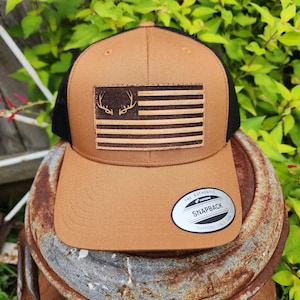 May include: A brown and black baseball cap with a leather American flag patch featuring a deer antler design. The cap has a black mesh back and a "The Authentic Yupoong Snapback" label.