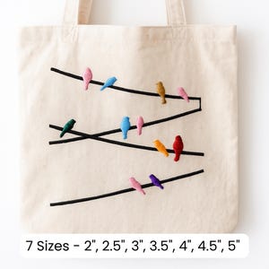 May include: Beige tote bag featuring an embroidered design of colorful birds perched on black lines. The birds are in various colors, including pink, blue, green, yellow, and purple. The text at the bottom reads "7 Sizes - 2", 2.5", 3", 3.5", 4", 4.5", 5""