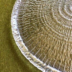 May include: A clear glass serving plate with a textured, rippled surface. The plate has a circular shape and a raised edge.