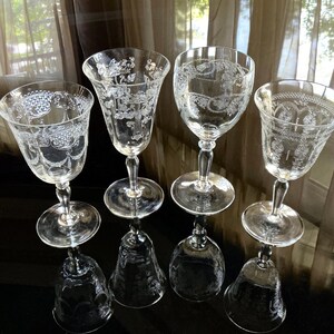 May include: Four clear, etched glass goblets with ornate floral and scroll designs. Each goblet has a round base, a slender stem, and a flared bowl. The glasses are arranged on a reflective surface, with their inverted reflections visible below.