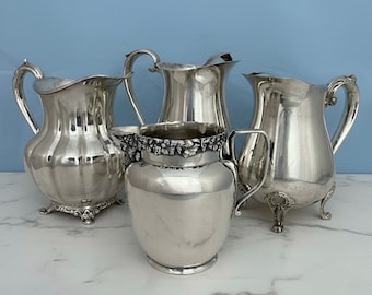 May include: A collection of four silver-toned metal pitchers. One pitcher features a prominent floral embossed rim, while the others display smooth, fluted, or ornate footed bases. Each vessel includes a curved handle and a pouring spout.