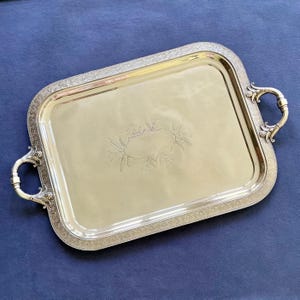 May include: A rectangular silver-toned serving tray with rounded corners and two ornate handles. The tray features a wide, decorative border with an intricate pattern and a central engraved design consisting of a blank cartouche with foliage.