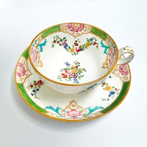 May include: A white teacup and saucer set with a floral design. The cup and saucer have a green border and gold trim. The floral pattern includes pink, yellow, and blue flowers. The cup has a curved handle.