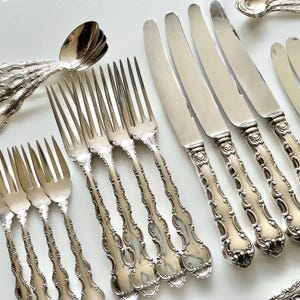 May include: An assortment of antique silverware, including forks, knives, and spoons, arranged on a white surface. The silverware features ornate, detailed handles and a polished, reflective finish, suggesting a vintage or heirloom quality. The set is ideal for formal dining.