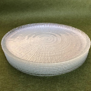 May include: A stack of eight clear glass plates with a textured, rippled design. The plates are stacked on a green surface.