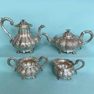 May include: A silver tea set with intricate floral patterns. The set comprises a teapot, a creamer jug, a sugar bowl, and a coffee pot. Each piece features curved handles and decorative feet. The background is a light blue.