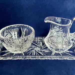 May include: A clear, cut-glass serving set with a creamer jug, a sugar bowl, and a rectangular tray. The set has a starburst pattern and is suitable for serving tea or coffee. The tray is approximately 25 cm long.