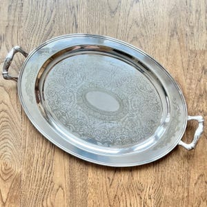 May include: Oval silver serving tray with ornate floral and scrollwork design. The tray has two curved handles and a polished finish. The centre of the tray features an oval design. A decorative piece for serving food or displaying items.
