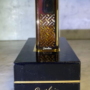 May include: A gold-toned perfume bottle with a geometric design on the front, resting on a black box with gold trim. The brand name "Guerlain" is visible on the box. The bottle has a sleek, rectangular shape.
