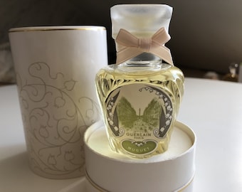 May include: A small bottle of Guerlain Paris Muguet perfume in a white box with a gold trim. The bottle is clear glass with a floral design and a white bow. The box has a floral pattern.