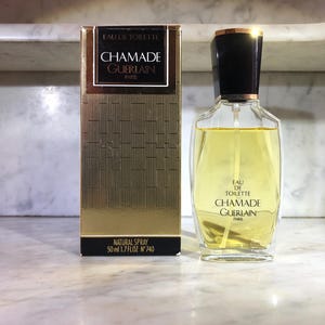 Könnte beinhalten: Eine goldene und schwarze Flasche Chamade Eau de Toilette von Guerlain. Die Flasche enthält 50 ml und ist ein natürlicher Sprühkopf. Die Flasche steht auf einer weißen Marmorfläche.