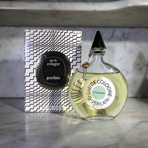 May include: A clear glass bottle of Guerlain Chamade Eau de Cologne. The bottle has a gold cap and a label with the text "Eau de Cologne Chamade Guerlain" in a circle.