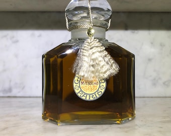 Può includere: Un flacone di profumo vintage con un tappo di vetro trasparente e un nappa decorativa. La bottiglia è riempita con un liquido marrone dorato e presenta un'etichetta con la scritta "Mitsouko Paris". La bottiglia ha una forma ottagonale.