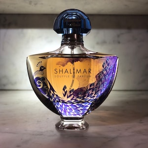 Könnte beinhalten: Eine goldene und blaue Glasflasche von Shalimar Souffle de Parfum von Guerlain Paris. Die Flasche zeigt ein Pfauenmotiv und den Text "Shalimar Souffle de Parfum".