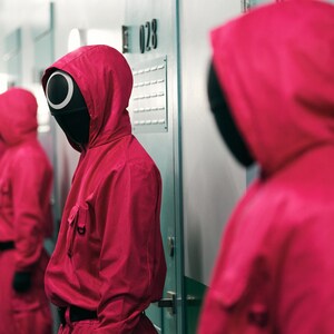 Op de afbeelding: Een groep mensen die rode jumpsuits en zwarte maskers dragen met een enkele grote cirkel voor een oog. De maskers zijn allemaal naar voren gericht. De mensen staan in een rij voor een muur met lockers.