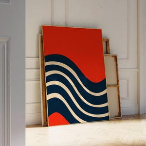 May include: A framed abstract art print featuring a vibrant red background with a navy blue and cream wave design. The artwork is leaning against a white wall, showcasing a modern aesthetic.