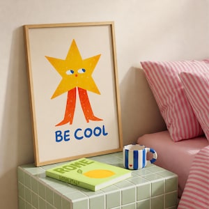 May include: A framed wall art print featuring a yellow star character with blue eyes and orange legs, with the text BE COOL in blue letters below. The print is displayed next to a book titled ROME ITALY and a blue and white striped ceramic mug.