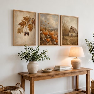 Op de afbeelding: Drie ingelijste wandkunstwerken op een rij. De linker print toont bruine eikenbladeren en eikels. De middelste print toont een pompoenveld met oranje pompoenen. De rechter print toont een rustieke schuur in een veld onder een bewolkte lucht.