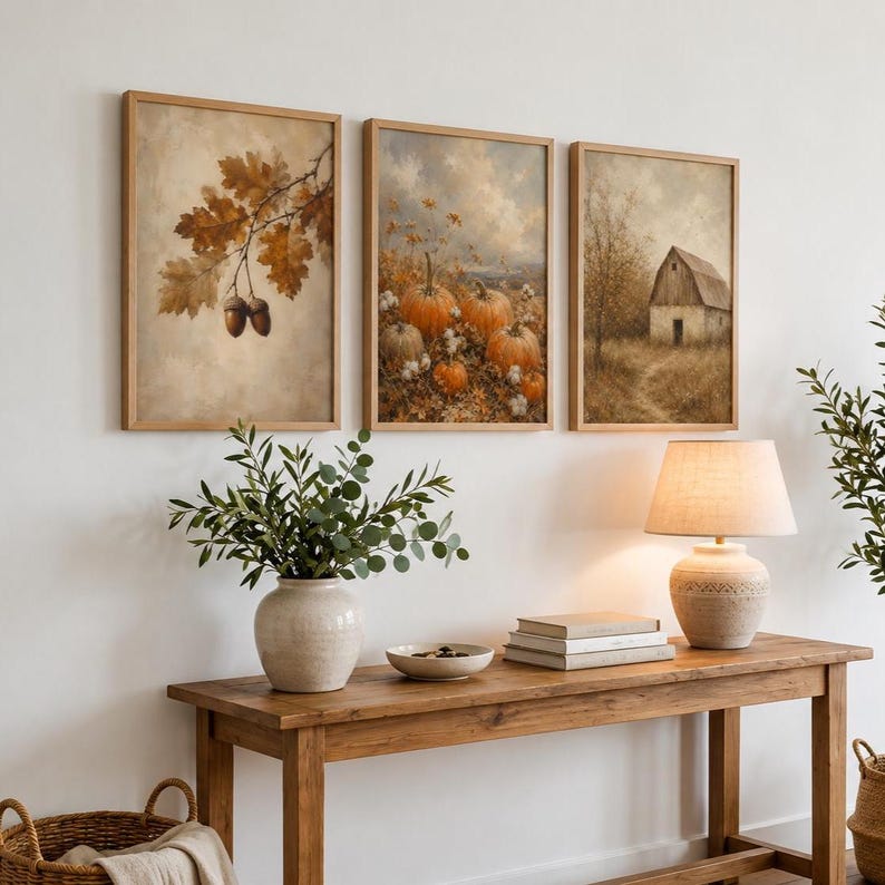 Op de afbeelding: Drie ingelijste wandkunstwerken op een rij. De linker print toont bruine eikenbladeren en eikels. De middelste print toont een pompoenveld met oranje pompoenen. De rechter print toont een rustieke schuur in een veld onder een bewolkte lucht.