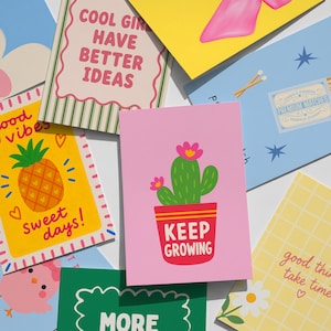 May include: A collection of colorful greeting cards featuring various illustrations and text. Designs include a green cactus in a red pot with the text KEEP GROWING, a yellow card with a pineapple and the text sweet days, and other cards with patterns, flowers, and phrases like COOL GIRL HAVE BETTER IDEAS.