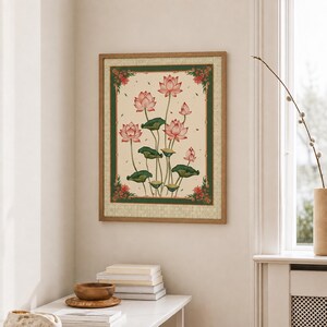May include: A framed wall art print featuring a botanical illustration of pink lotus flowers with green leaves on a cream background, enclosed by a dark green border with small red floral accents at each corner, set in a light wood frame.