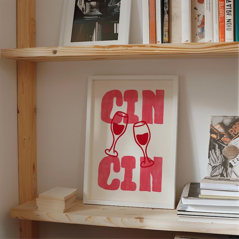 May include: A framed rectangular print featuring the text CIN CIN in bold pink letters, with two red wine glasses illustrated in the center, positioned between the words. The artwork is displayed in a simple white frame on a wooden shelf.