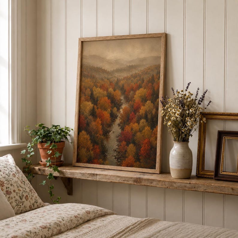 Op de afbeelding: Een ingelijste verticale wandkunstprint met een landschap van een kronkelende rivier door een dicht bos met herfstbladeren in tinten oranje, rood en geel onder een zachte, bewolkte lucht, geplaatst op een houten plank.