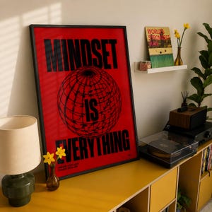 K&ouml;nnte beinhalten: Ein gerahmtes rotes Poster mit den Worten "MINDSET IS EVERYTHING" in Schwarz und einer Weltkugel-Grafik. Das Poster steht neben einem gelben Schrank mit Lampe und Plattenspieler.