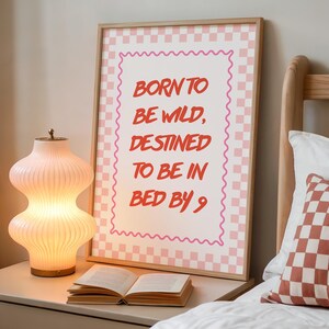 May include: A rectangular framed wall art print featuring a pink and white checkered border and a wavy pink frame line. The center text in red reads: BORN TO BE WILD, DESTINED TO BE IN BED BY 9. The print is displayed in a light wood frame.