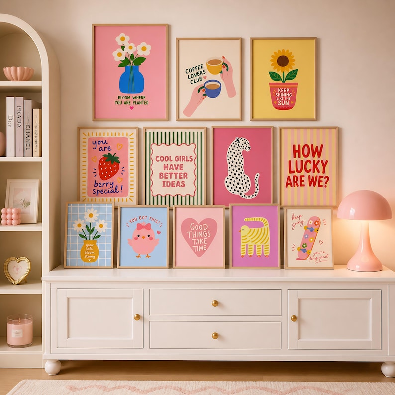 May include: A collection of twelve framed wall art prints featuring colorful illustrations and text, including a strawberry, a leopard, a sunflower, and various motivational phrases, displayed on a white wooden sideboard with a pink lamp.