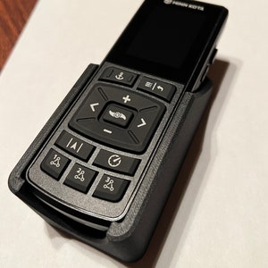 Op de afbeelding: Een zwarte afstandsbediening met een grijze kunststof behuizing en een klein scherm. De afstandsbediening heeft verschillende knoppen, waaronder een directionele pad, een plus- en min-knop en een knop met een anker symbool. De tekst "Minn Kota" is gedrukt op de bovenkant van de afstandsbediening.