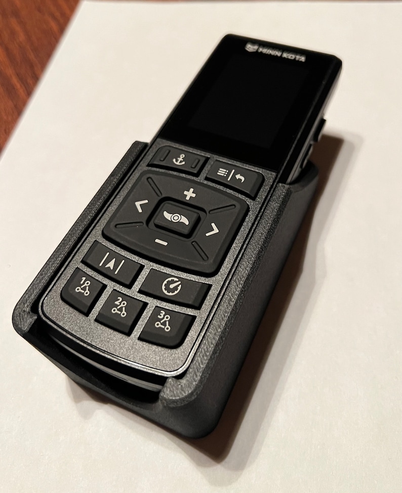 Op de afbeelding: Een zwarte afstandsbediening met een grijze kunststof behuizing en een klein scherm. De afstandsbediening heeft verschillende knoppen, waaronder een directionele pad, een plus- en min-knop en een knop met een anker symbool. De tekst "Minn Kota" is gedrukt op de bovenkant van de afstandsbediening.