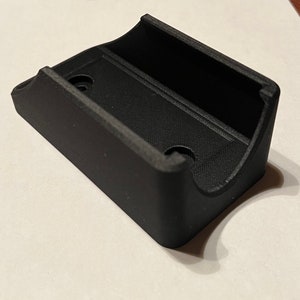 Op de afbeelding: Een zwarte plastic wandbevestiging met twee gaten voor bevestigingsschroeven. De bevestiging heeft een gebogen vorm om een cilindrisch voorwerp vast te houden.