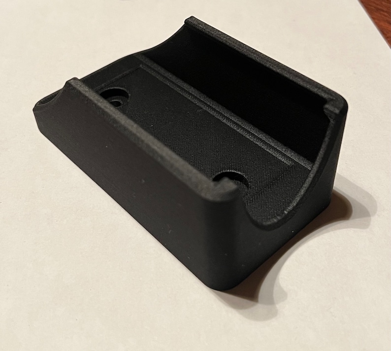 Op de afbeelding: Een zwarte plastic wandbevestiging met twee gaten voor bevestigingsschroeven. De bevestiging heeft een gebogen vorm om een cilindrisch voorwerp vast te houden.