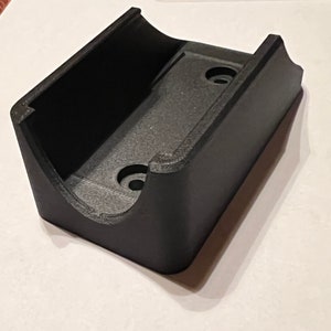 Op de afbeelding: Zwarte plastic wandbeugel met twee schroefgaten. De beugel heeft een gebogen vorm om een object vast te houden.