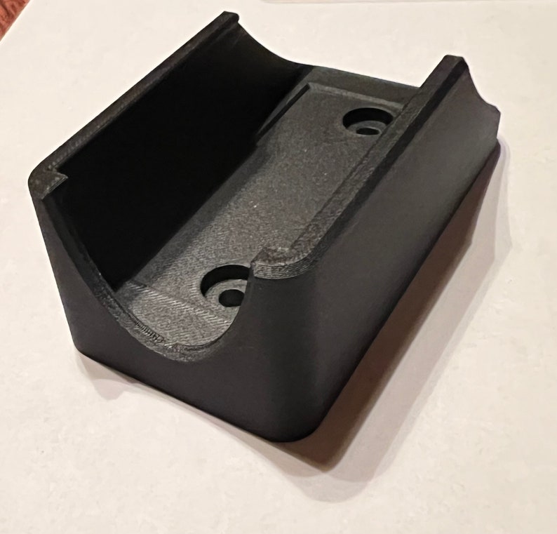 Op de afbeelding: Zwarte plastic wandbeugel met twee schroefgaten. De beugel heeft een gebogen vorm om een object vast te houden.