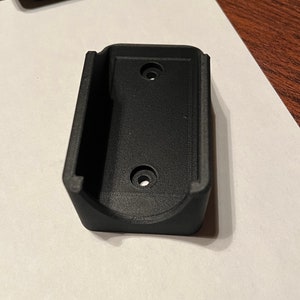 Op de afbeelding: Een zwarte plastic wandbeugel met twee gaten voor schroeven. De beugel is ontworpen om een klein elektronisch apparaat te houden.