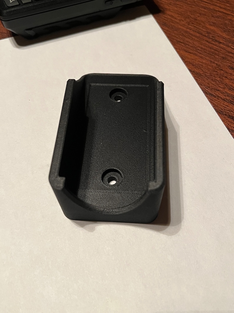 Op de afbeelding: Een zwarte plastic wandbeugel met twee gaten voor schroeven. De beugel is ontworpen om een klein elektronisch apparaat te houden.