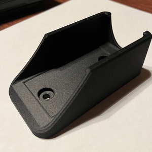 Op de afbeelding: Een zwarte plastic wandbeugel met twee gaten voor bevestigingsschroeven.