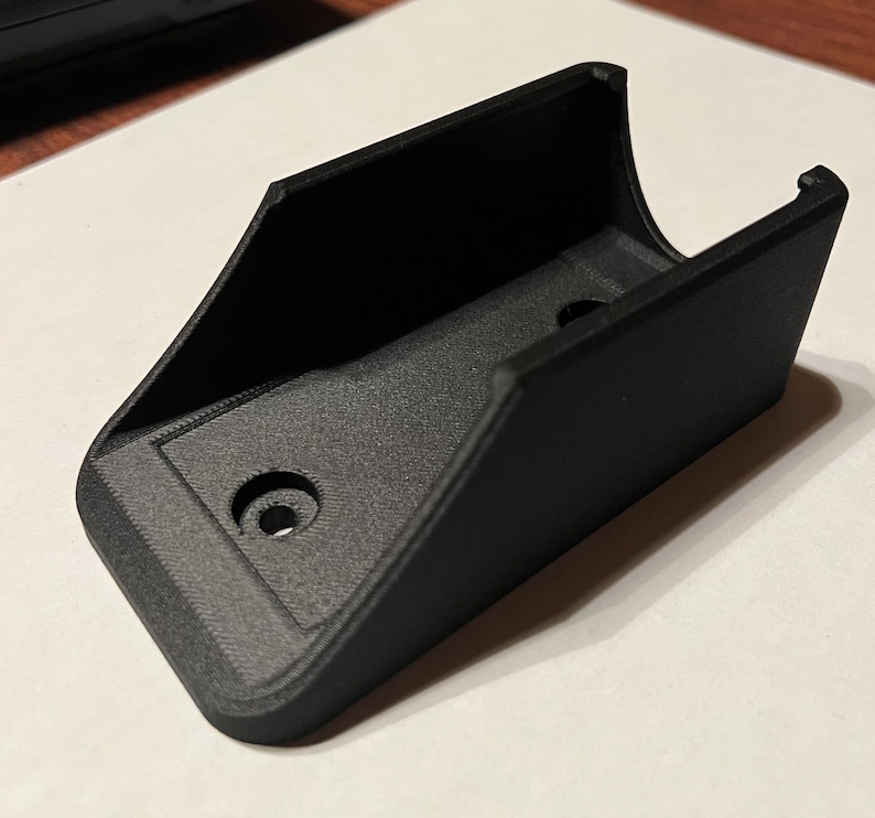 Op de afbeelding: Een zwarte plastic wandbeugel met twee gaten voor bevestigingsschroeven.