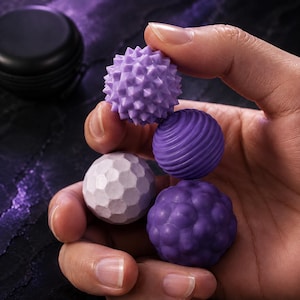 Op de afbeelding: Een set van vier kleine, bolvormige massageballen in verschillende tinten paars en lavendel. Elke bal heeft een eigen oppervlaktestructuur, waaronder scherpe punten, horizontale ribbels, een gefacetteerd patroon en afgeronde bobbels.
