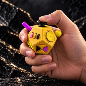 Puede incluir: Un juguete antiestrés amarillo de varias caras con un joystick negro en la parte superior, un dial circular amarillo, una pequeña esfera amarilla y un interruptor morado. El dispositivo incluye tres pequeñas ruedas giratorias en rosa, amarillo y negro.
