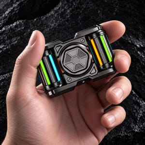 Könnte beinhalten: Ein rechteckiges Fidget-Spielzeug aus dunkelgrauem Metall mit einer zentralen runden Platte mit geometrischem Muster und zwei zylindrischen Seitenelementen. Jeder Zylinder zeigt vertikale Lichtstreifen in leuchtendem Orange, Grün und Blau.
