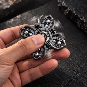 May include: A dark gray metal fidget spinner with a complex, multi-armed design featuring exposed silver ball bearings in each section. The device is captured in motion, showing a blurred spinning effect around its central axis and frame.